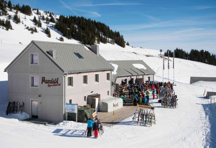 Alprestaurant Panüöl restaurant surrounded by snow, with a hill behind it, and lots of people in ski suits sitting on the terrace