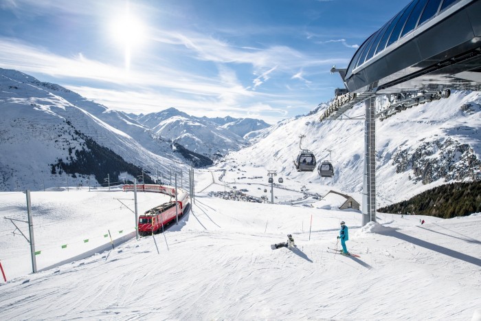 Two skiiers on a slope at Andermatt, watching a red train pass uphill near them