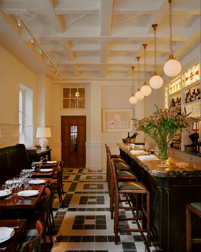 The hotel’s Brasserie l’Émil, with a row of darkwood tables and chairs, a black and white tiled floor and seats against a marble counter over which hangs a row of globe lamps