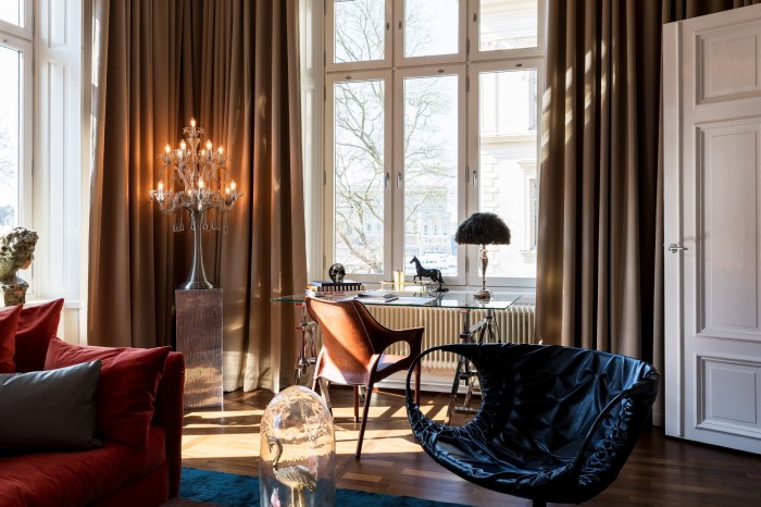 A room in Stockholm’s Lydmar Hotel, with a red velvet sofa, modernist black leather seat, ceiling to floor brown curtains and a chandelier-style lamp perched on a tall metallic stand