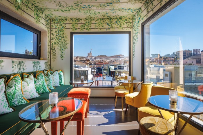 A bar space in the Singer Palace Hotel with large windows overlooking central Rome, green, yellow and orange velvet chairs, stools and banquette and walls covered with a trompe l’oeil of ivy