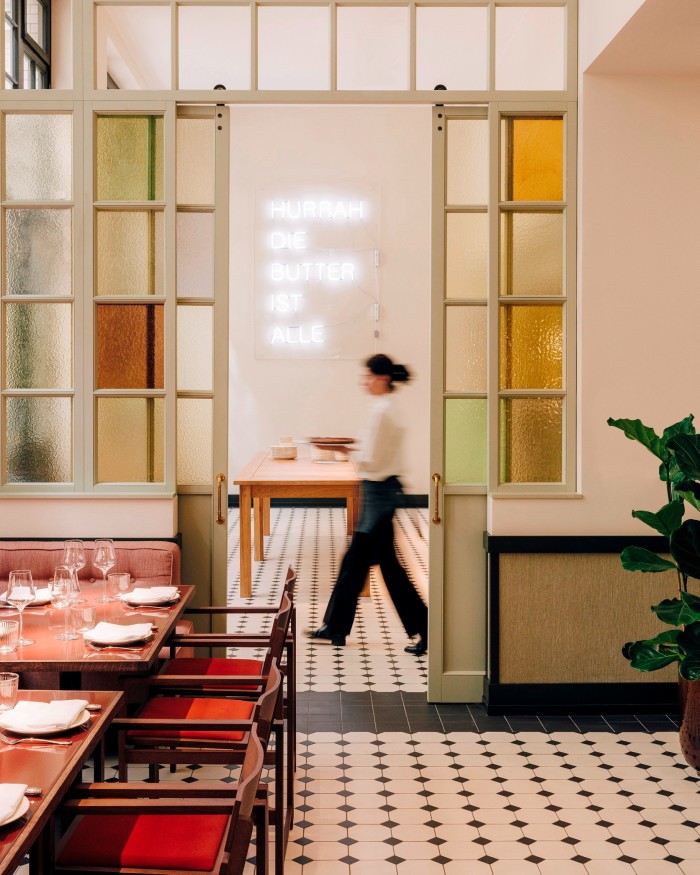 A detail of the Château Royal’s mid-century style restaurant, with a woman holding a plate walking past a white neon artwork that reads “Hurrah, Die Butter Ist Alle” 