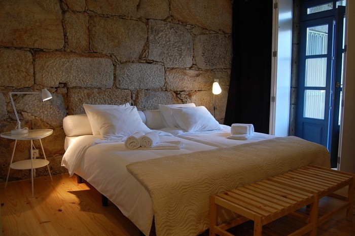 A bed in a room in Casa dos Guindais, with a pale-wood floor and a wall made of up  of large, irregular soft-brown bricks