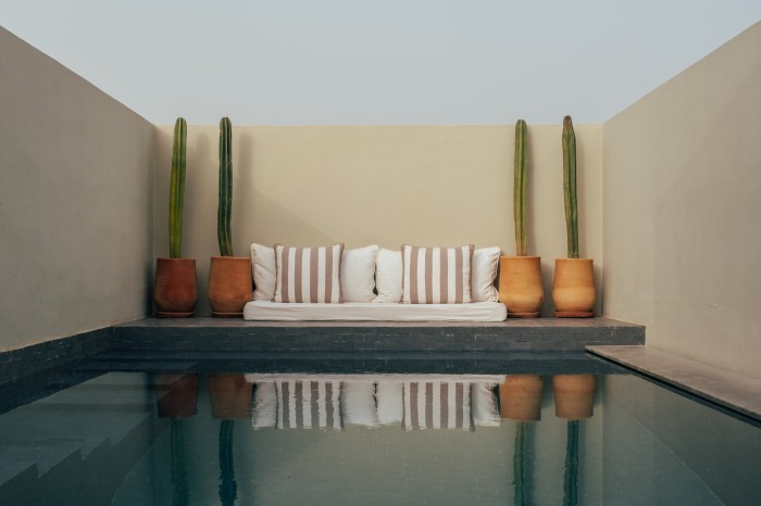 The open-air pool at Riad No 37, surrounded by smooth pale beige walls and with large cacti in terracotta pots flanking large cushions 