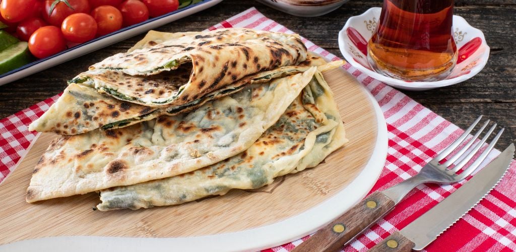 on a wooden table, there is a pancake Gözleme with cheese and herbs. A cherry tomato, tea, and a fork and bread knife 