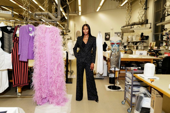 Naomi Campbell wearing a black suit in a fashion workroom, standing beside a long fluffy pink robe