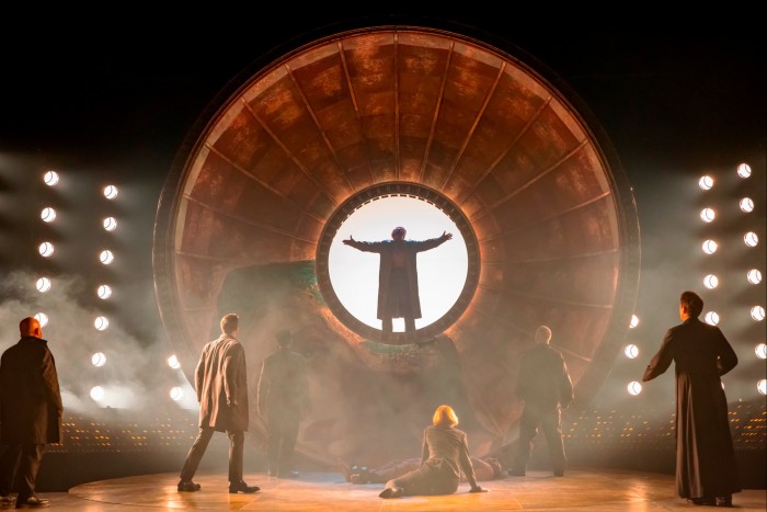 A male singer standing inside a giant wheel watched by four male members of the cast on a smoky stage in ‘Tosca’