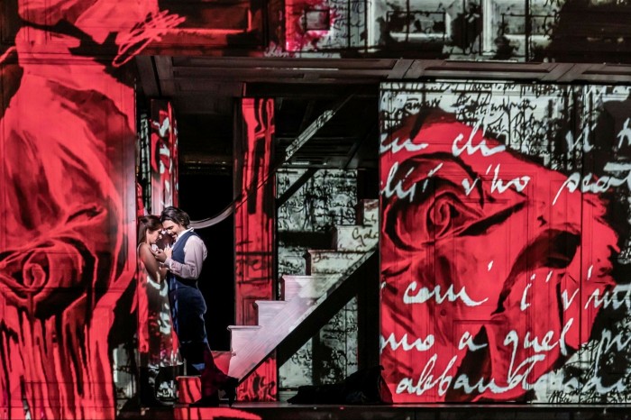 A scene from the Canadian Opera Company’s ‘Don Pasquale’: a man and a woman standing at the bottom of a staircase, on either side of which are walls covered in graffiti and paintings of red roses