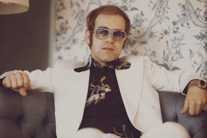 Elton John seated in a chair wearing a white blazer and sunglasses