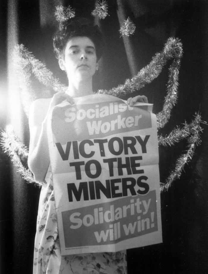 ‘Support the Miners: Solidarity Will Win!’, 1984, by Alison Lloyd: a black and white photograph of a woman holding a Socialist Worker poster that reads ‘Victory to the Miners’
