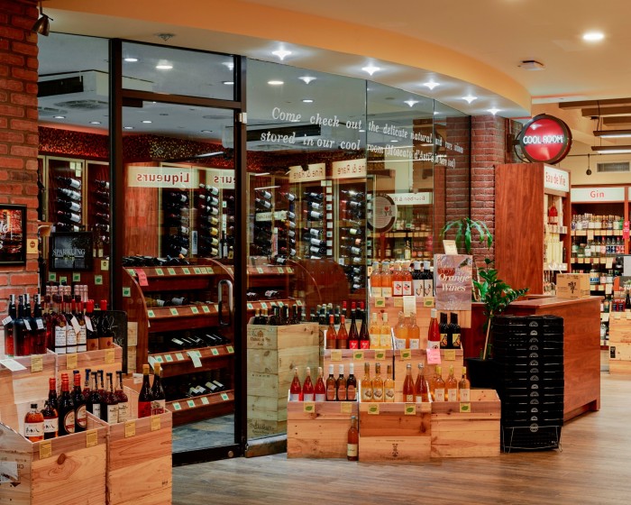 The interior of Astor Wines & Spirits, with a large glass front door, small wooden crates and a barrel on which stand bottles of wine, and shelves stacked with wine bottles