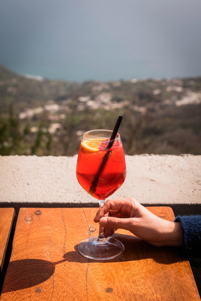 A person holds a glass at a table, with a view to the sea behind 