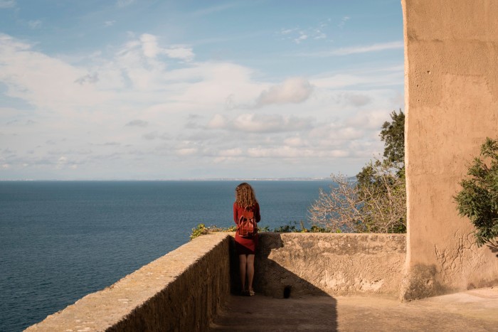 A woman with a backpack, seen from behind, gazes out from castle walls to the sea