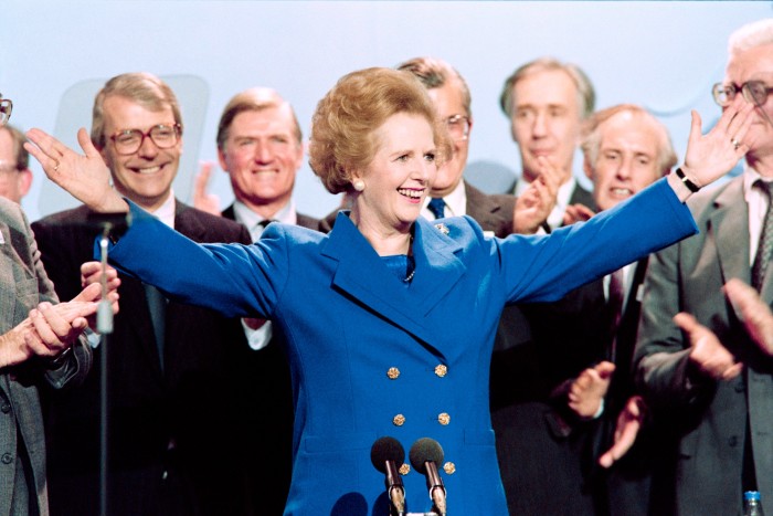 Margaret Thatcher, flanked by applauding men, smiles and spreads her arms wide