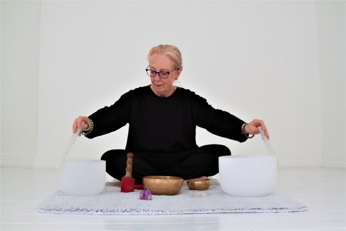 Jane Kight, a bespectacled woman dressed in black, sitting cross-legged as she rotates rods around the rims of two white singing bowls