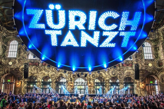 Hundreds of people beneath a giant blue sign that says ‘Zürich Tanzt’ in the city’s main train station