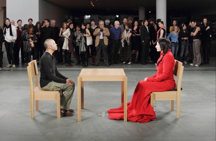 Marina Abramović in a long red dress sitting at a table in her ‘The Artist Is Present’ piece at New York’s Museum of Modern Art, 2010. A man with a shaved head is sitting opposite her and spectators are watching from a distance