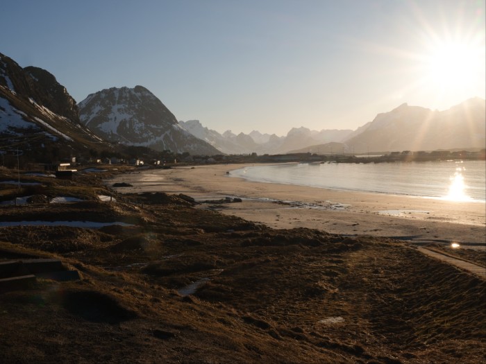 Sunlight bursts from behind a mountain on the right. On the left and in the foreground, rocky slopes reach down to the shore 