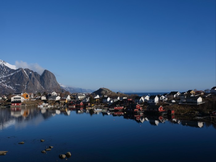 The houses of a coastal village strung along the shoreline under a blue sky reflected in still blue water with snow-streaked mountains in the background