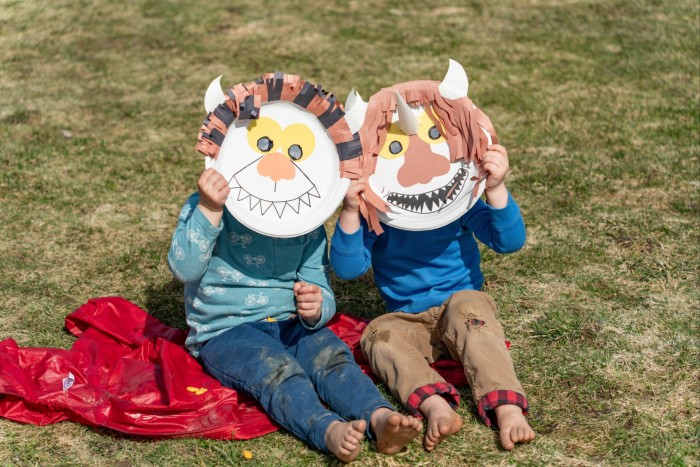 Two young children sit on grass, holding monster-face viewing masks up to their faces