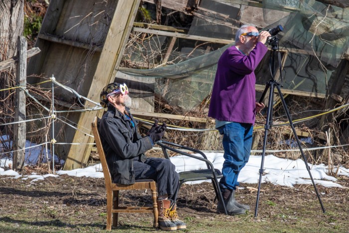 One man stands next to a camera tripod, near another man sitting on a chair. Both look to the sky through dark glasses