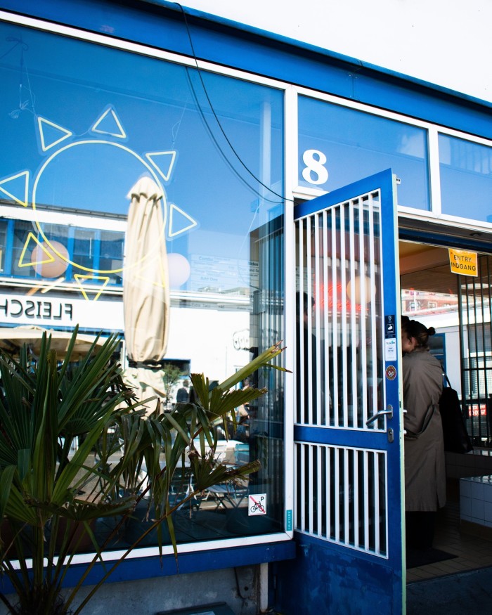 The facade of Hija de Sanchez taqueria, with its blue-framed door and window, in which hangs a yellow neon installation of the sun and a closed parasol and a white building are both reflected, as well as a blue sky