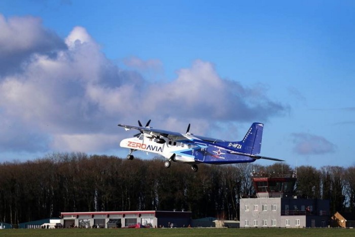 A propeller-driven aircraft with the inscription ‘ZEROAVIA’ is seen ascending above a grassy airfield with buildings and trees in the background