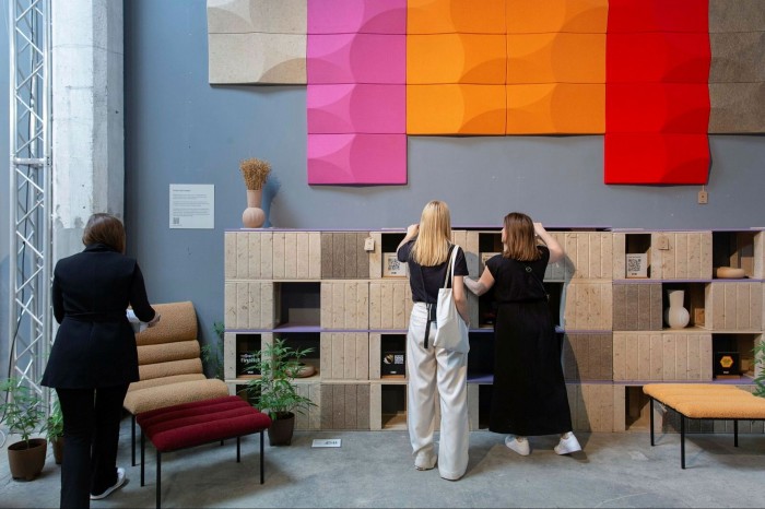 ‘Is One Life Enough?’, an exhibition at the Isola Design Festival: two women looking at brown, brick-like Modernist shelving, and another woman looking at a red and beige velvet Modernist chair. On the walls are large, multi-coloured, 3D tiles