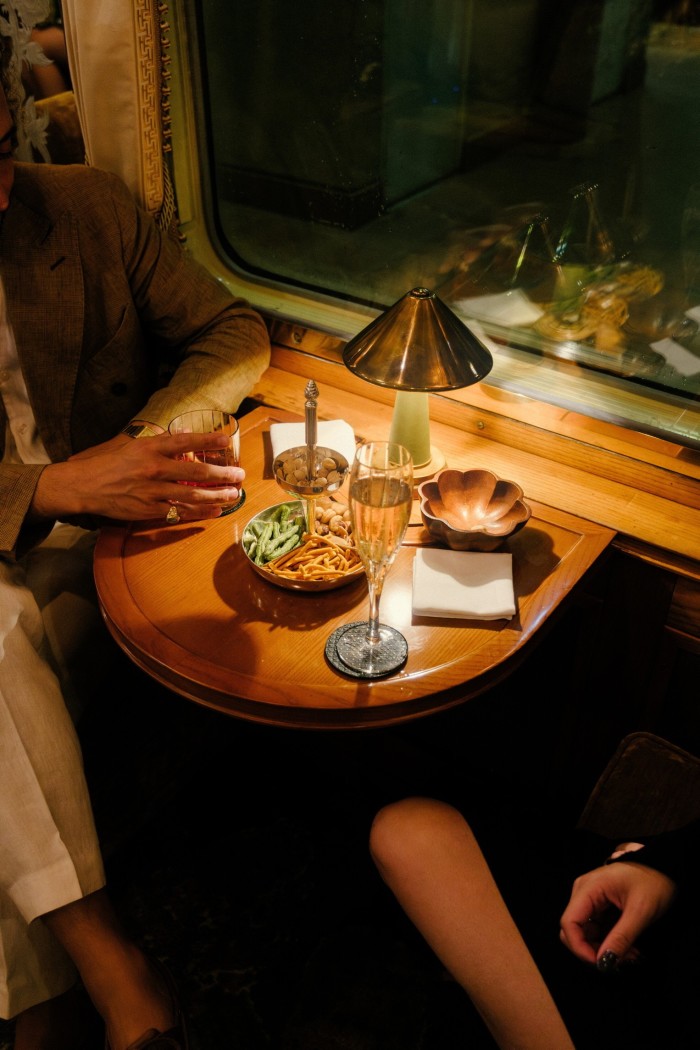 In the evening, with dark outside, two people sit at a small table where there are drinks and snacks