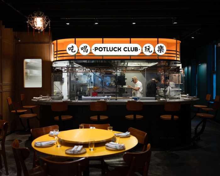 A circular table in front of Potluck Club’s open kitchen, with a white sign above it that reads ‘Potluck Club’ on an orange background 