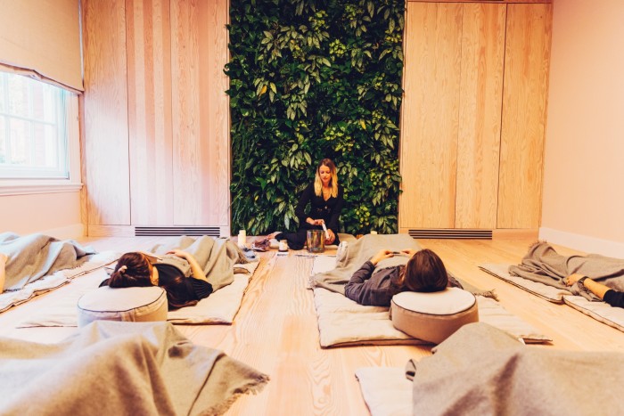 Attendees of a sound-bathing class at London’s Re:Mind Studio lying on the floor covered in linen blankets as a woman in the background rotates a wand around the rim of a singing bowl. Behind her, on the wall, is a green foliage installation