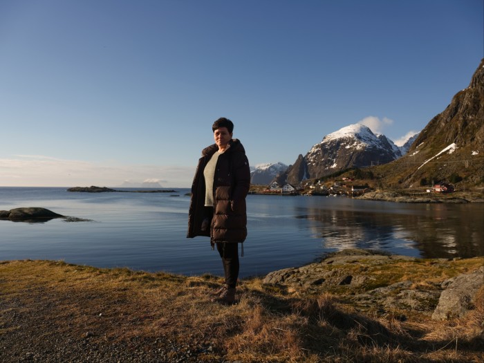 A woman in a long brown quilted winter coat stands on a grassy shore with water, blue sky and snowy peaks behind