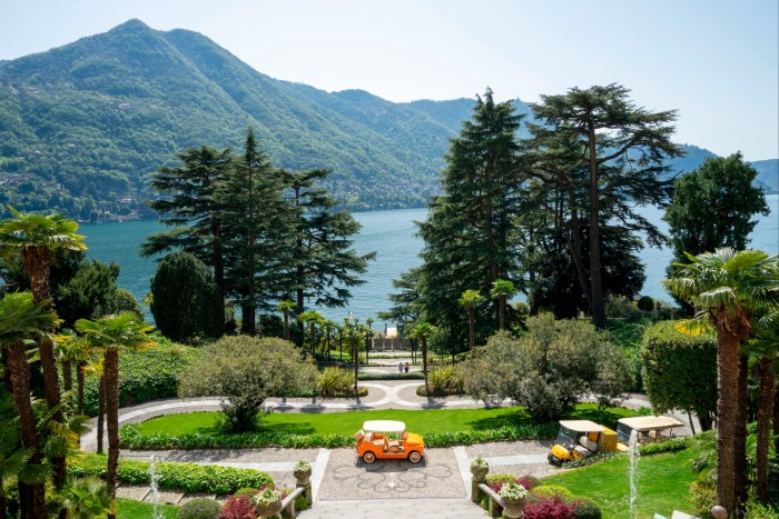 An orange car is parked next to a lawn on a terrace, with trees, a lake and hillsides beyond 