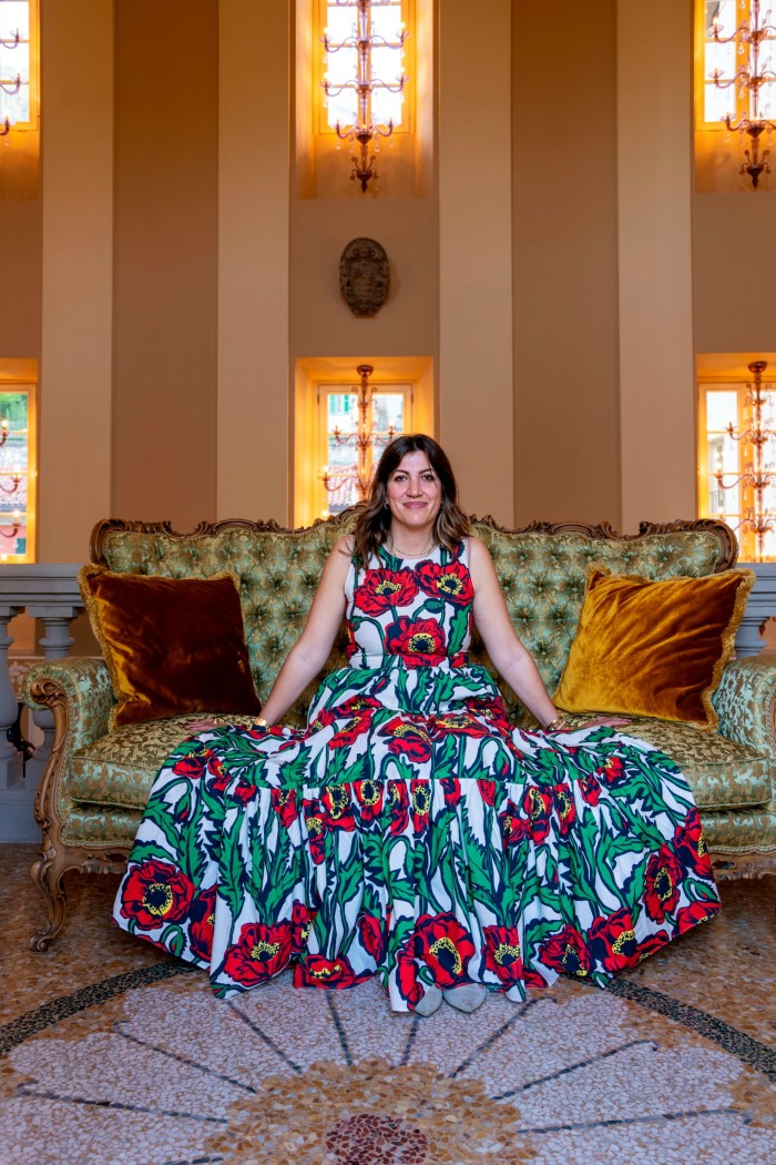 A dark-haired woman sits smiling on a sofa, the skirts of her long dress spread wide