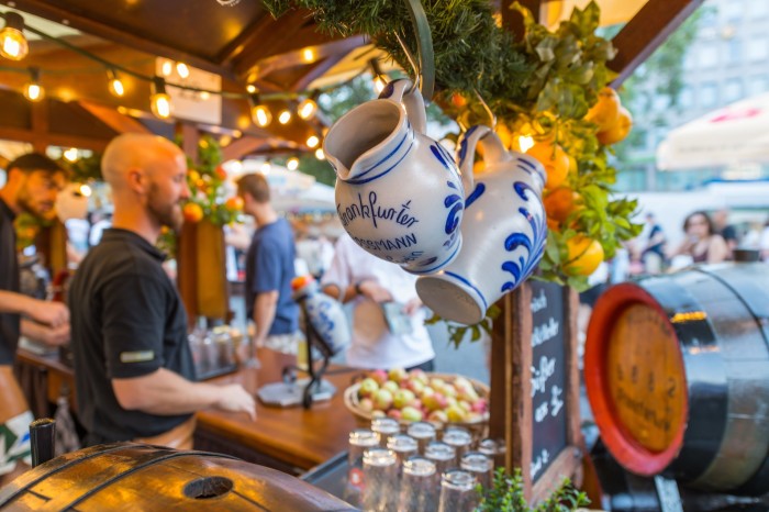 Two small blue and white jugs hanging from a stall at Frankfurt’s Apple Wine Festival, with two men and trays of glasses and apples in the background