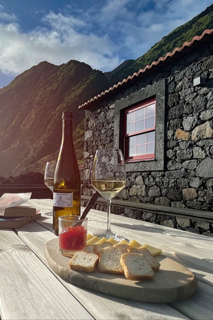 A bottle of wine, a cheeseboard and two wine glasses on a table. In the background are the wall of a house, a blue sky and a partial view of a mountain