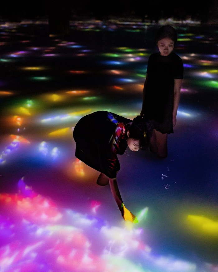 A woman watching another woman reach down to touch the digital koi in a teamLab Planets installation