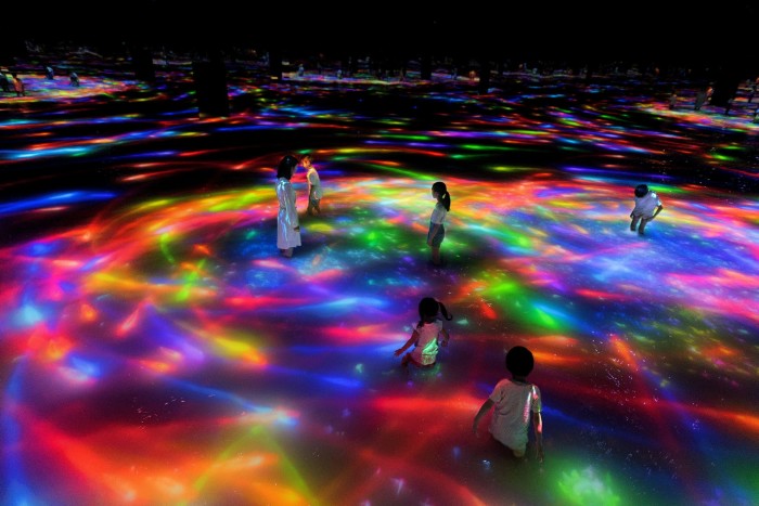 Adults and children standing knee-deep in water as digital koi in neon colours swim around them in a teamLab Planets installation