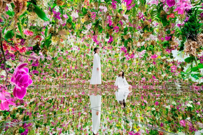 Two women – one standing up, the other kneeling on a mirrored floor – in teamLab’s Floating Flower Garden, a space filled with purple and white orchids