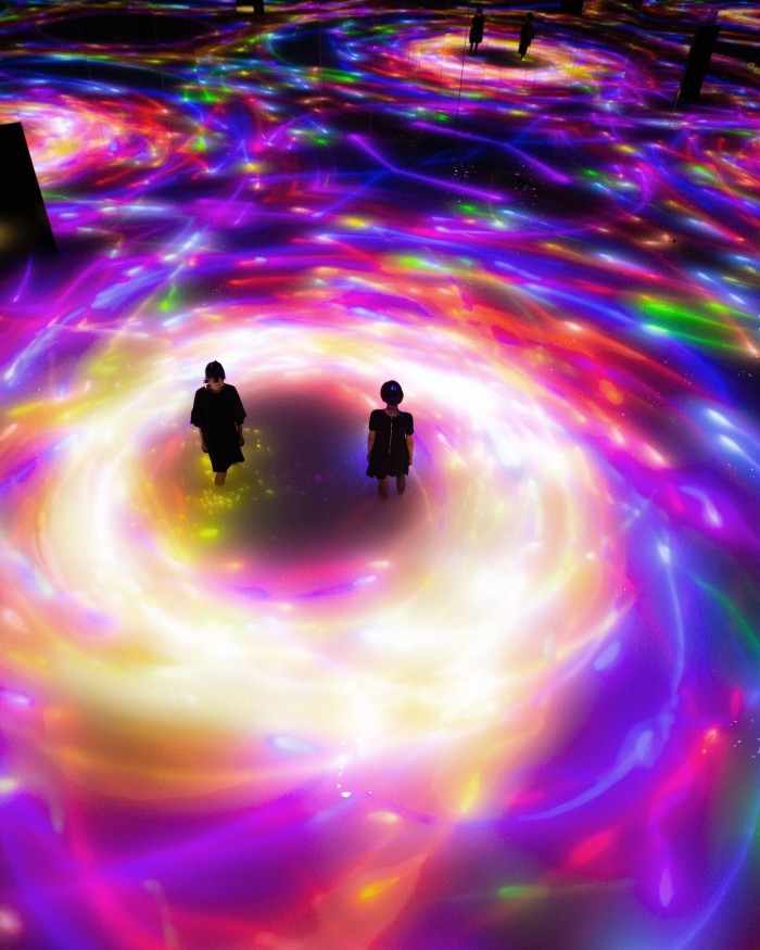 Two women photographed from above as a shoal of digital koi move around them in a yellow swirl in a teamLab Planets installation