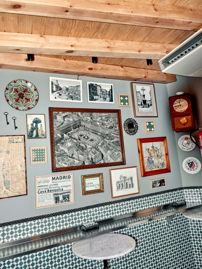Black and white photos and illustrations on the grey wall of Casa Revuelta; beneath them is a tiled lower wall, around which runs a zinc bar, and a small, circular marble-topped table
