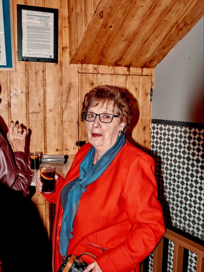 A bespectacled woman wearing a red jacket and blue silk scarf holding a drink at Casa Revuelta