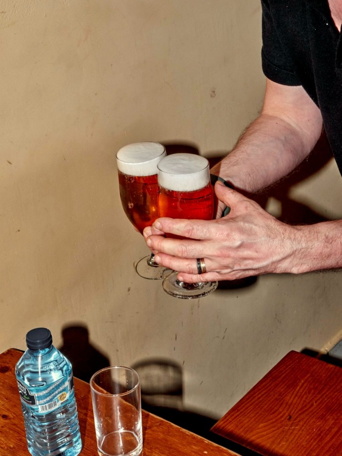  A man’s hands holding two glasses of beer above a wooden table on which stand a bottle of water and a glass at Casa Toni