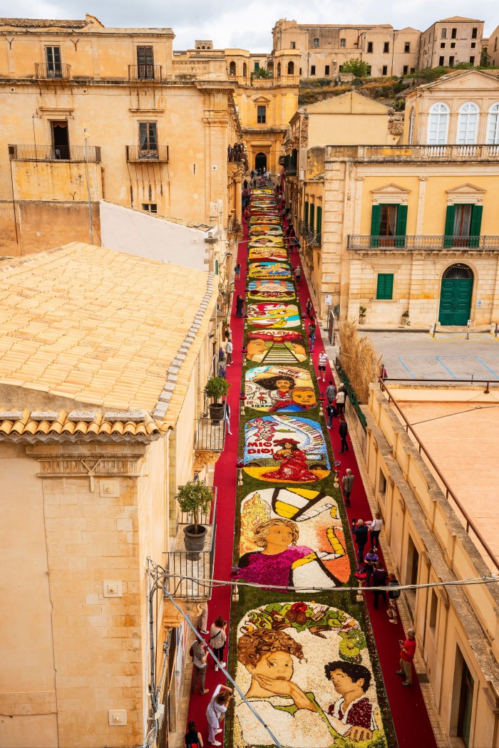 A narrow Italian street of historic sand-coloured baroque buildings lined for its entire length with floral tapestries