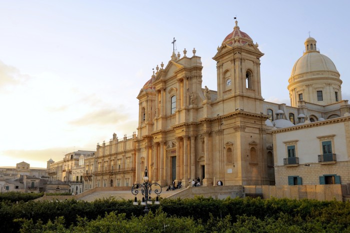 The ornate baroque facade of an Italian cathedral built from creamy coloured sandstone, with pillars, belltower and dome