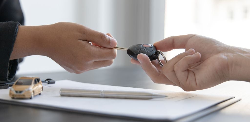 car rental agreement, sales rep handing over the keys to a car, paperwork on desk and small model car and pen on paperwork documents