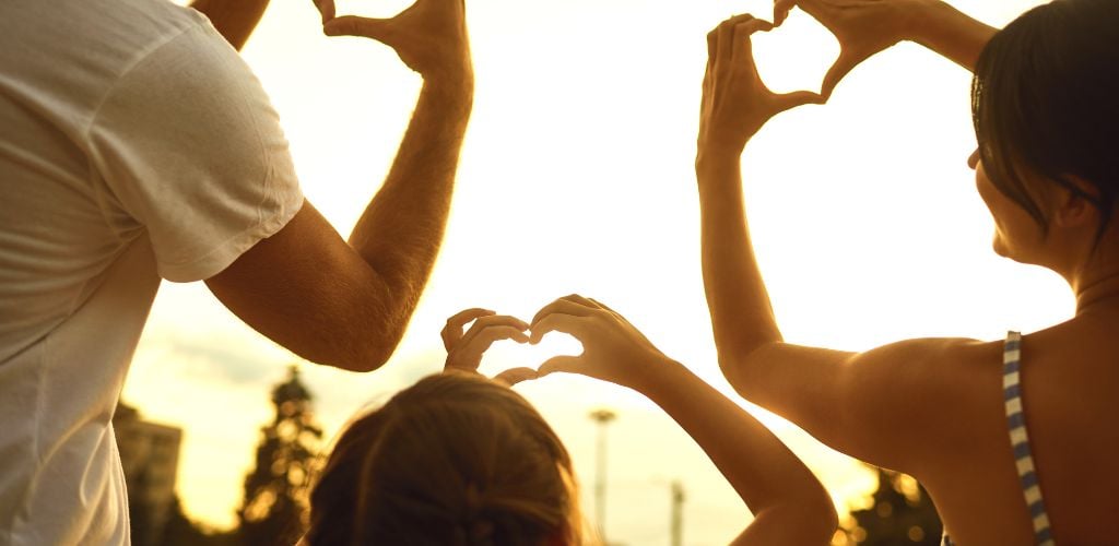family making heart shapes with their hands holding up against a sunset sky 