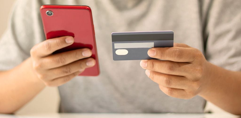 person holding smartphone in one hand and payment card in the other, wearing grey shirt 