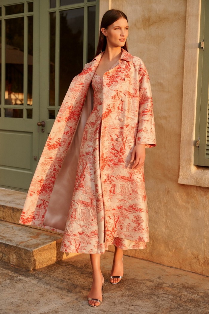 A model stands on a sunny street wearing a brocade dress and matching coat decorated with peach and cream coloured 18th-century landscape scenes 