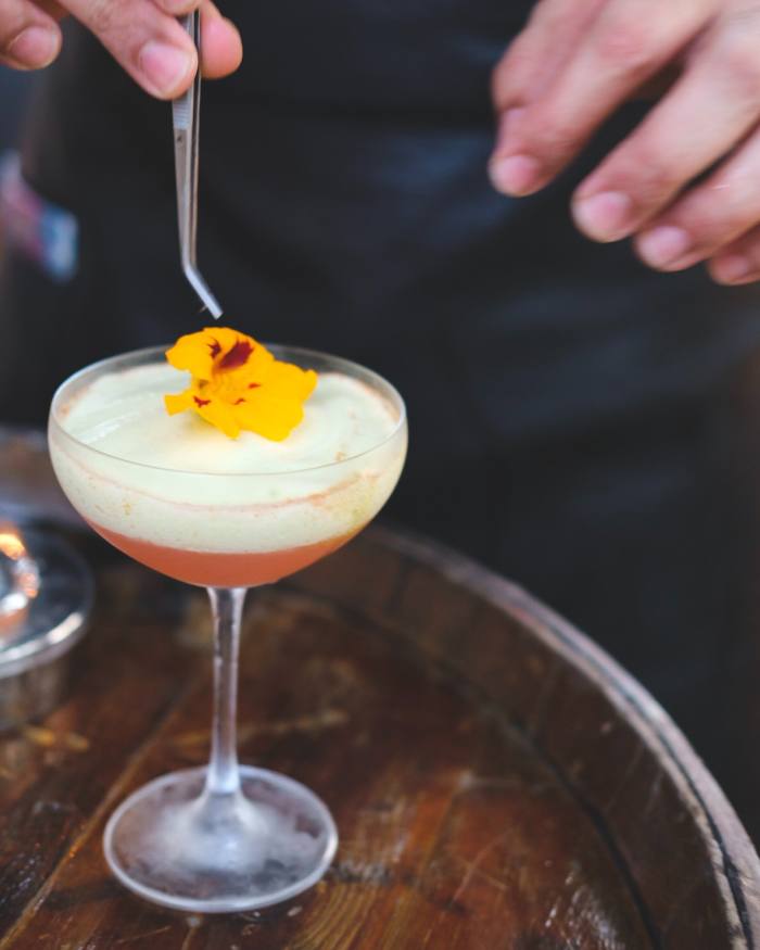 A cocktail with an orange-pink base, creamy top and a petal sitting on the top at COA bar